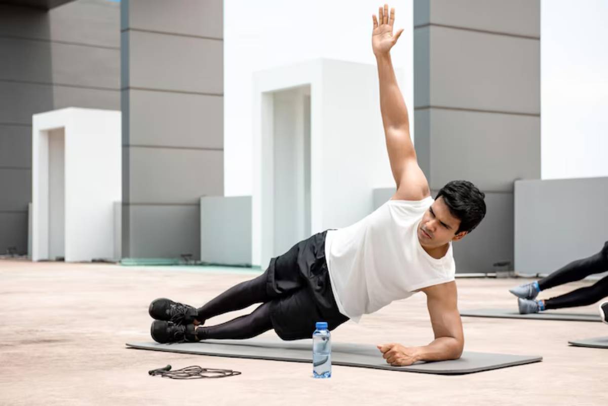 A man performs a side plank exercise on a yoga mat outdoors, with a water bottle and resistance bands nearby.