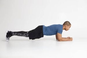 A person in a blue shirt and black shorts performs a plank on a white background, demonstrating core strength and stability.