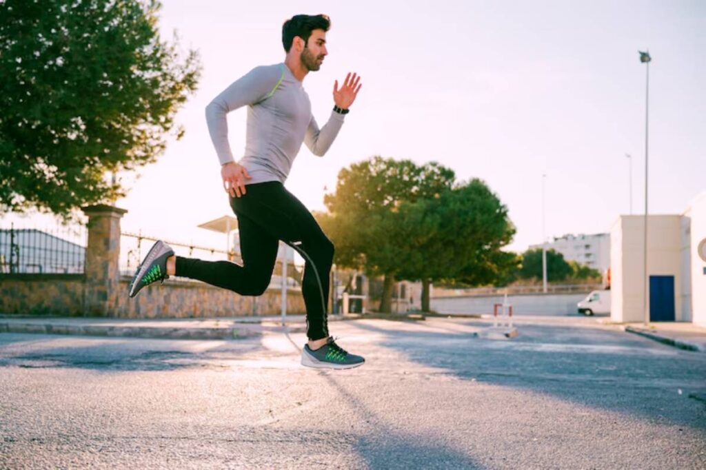 A man runs on a sunlit street, wearing athletic clothing and shoes, with trees and buildings in the background.