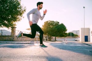 A man runs on a sunlit street, wearing athletic clothing and shoes, with trees and buildings in the background.