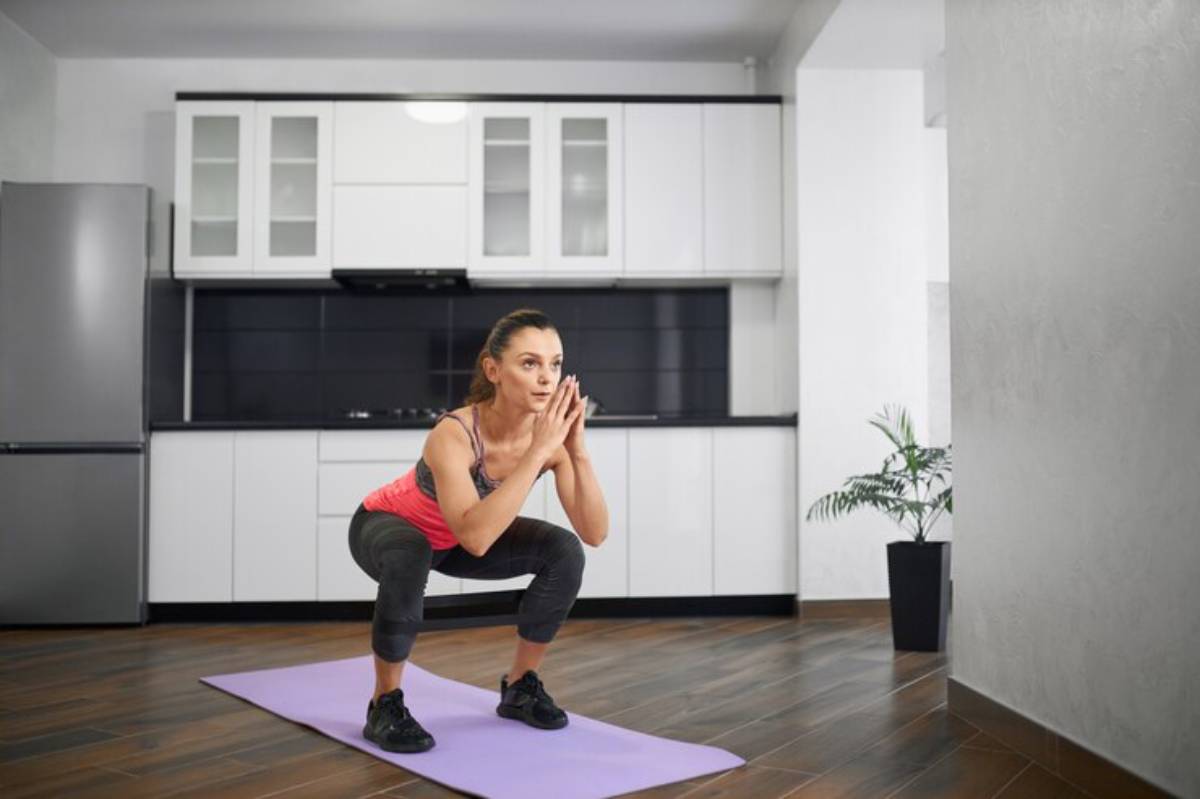 A woman performs a squat on a purple exercise mat in a modern kitchen with white cabinets and a potted plant in the background.