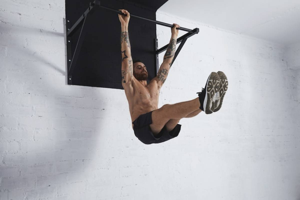 A shirtless individual with tattoos performs an ab exercise, gripping a pull-up bar while raising their legs.