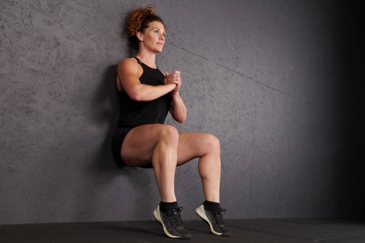 A person performs a wall sit exercise, squatting with legs parallel to the ground against a textured gray wall.
