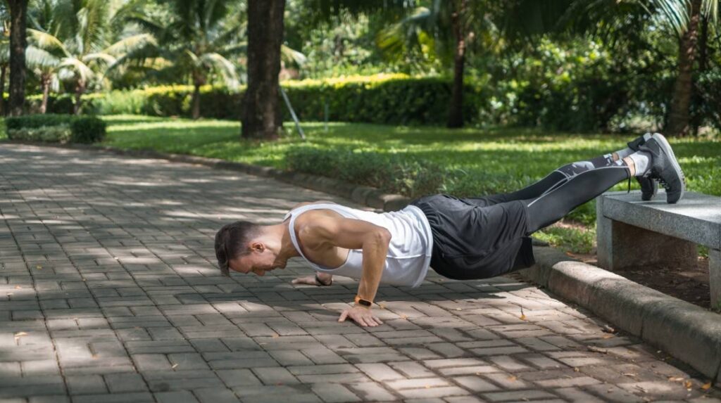 A person is performing a push-up on a stone bench in a park, surrounded by lush greenery and dappled sunlight.