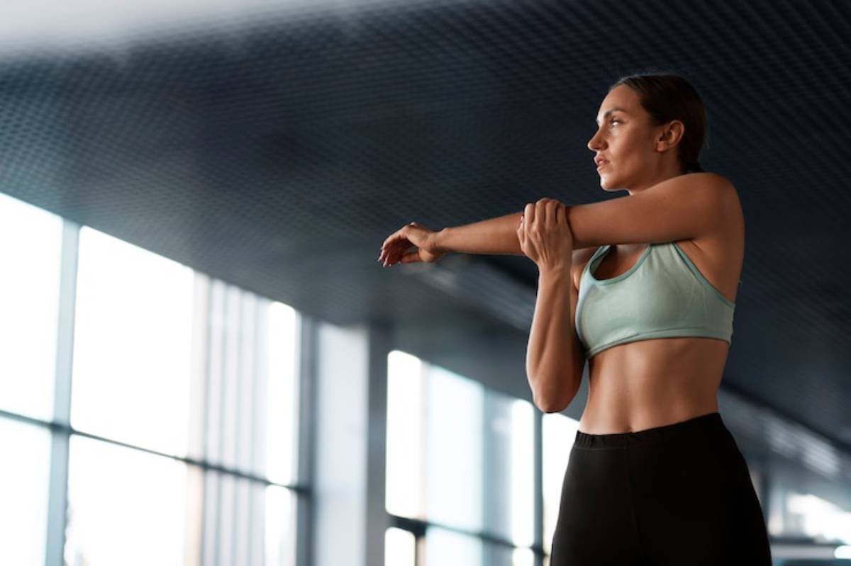 A fit woman stretches her arms, dressed in a teal sports bra and black leggings, against a backdrop of large windows.