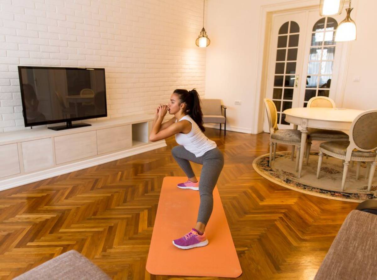 A woman performs squats on an orange exercise mat in a stylish, well-lit living room with wooden flooring and modern furniture.