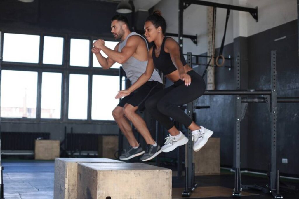 A man and a woman jump off a box in a gym, showcasing their fitness in a bright, spacious workout environment.