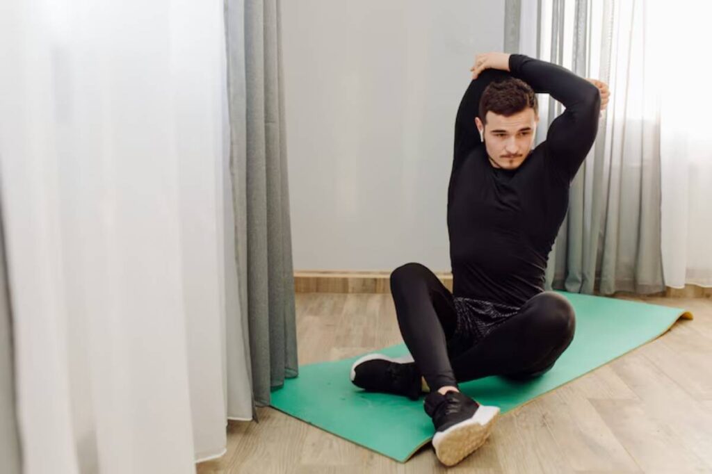 A person in activewear is seated on an exercise mat, performing a stretching exercise with one arm raised overhead.