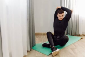 A person in activewear is seated on an exercise mat, performing a stretching exercise with one arm raised overhead.