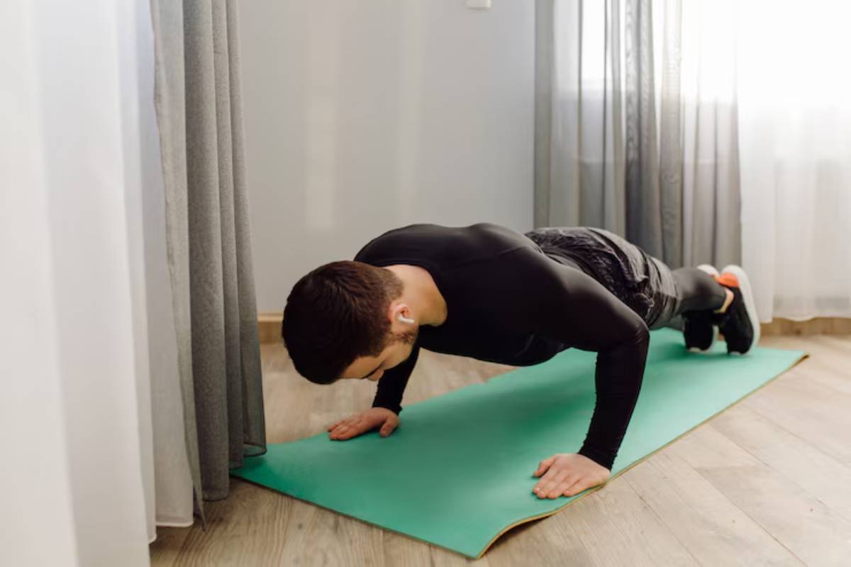 A man in a black athletic outfit doing push-ups on a green exercise mat in a well-lit indoor space.