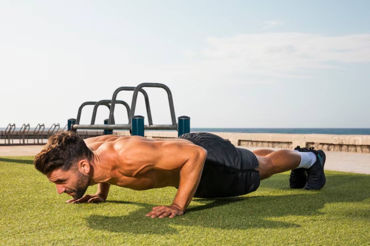 A muscular man performs push-ups on green turf, with outdoor fitness equipment and ocean views in the background under a clear sky.