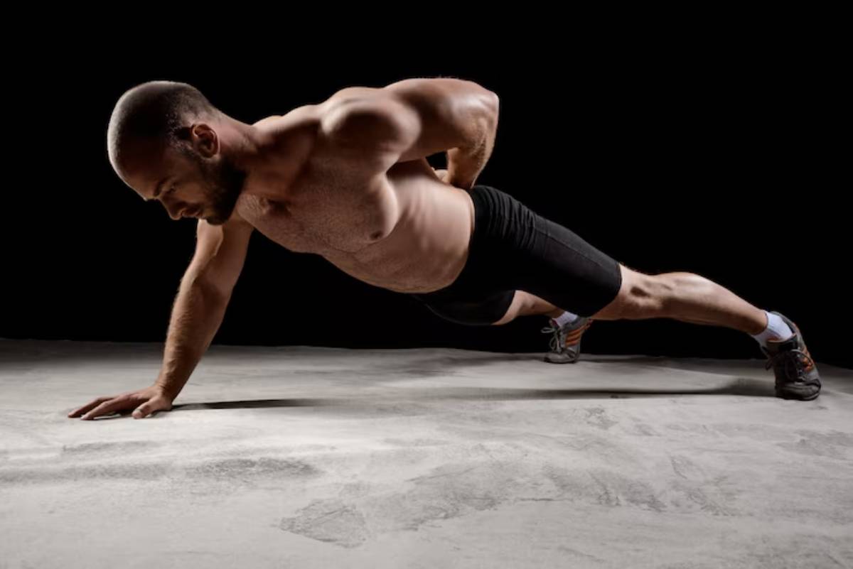 A muscular man performs a one-handed push-up on a concrete floor, showcasing strength and fitness against a dark backdrop.