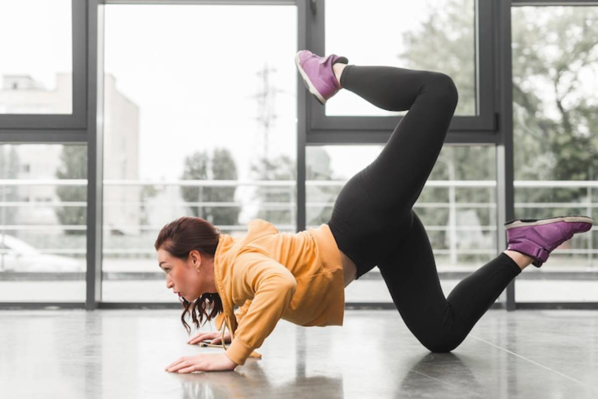 A person performs a dynamic exercise on a gym floor, wearing a yellow hoodie and black leggings, demonstrating strength and flexibility.
