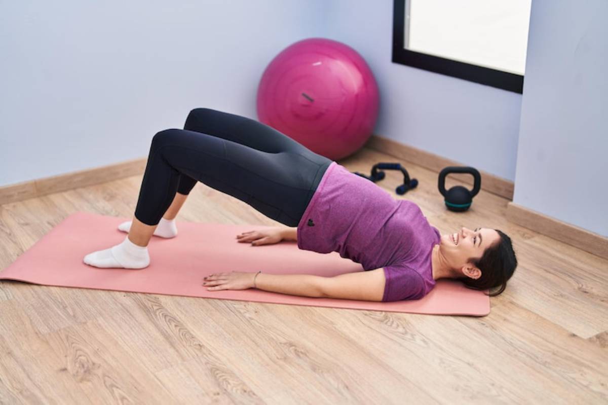 A person performs a bridge exercise on a pink yoga mat in a fitness room, with a stability ball and weights in the background.