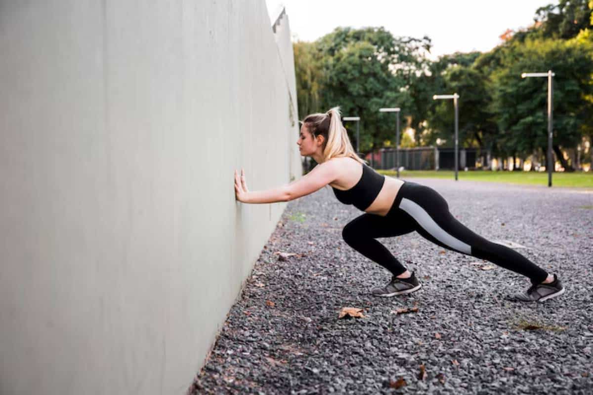 A woman in activewear performs an exercise against a wall in a park, surrounded by trees and soft gravel ground.